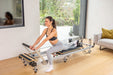 Woman exercising on a foldable metal reformer pilates machine with black upholstery in a home setting.
