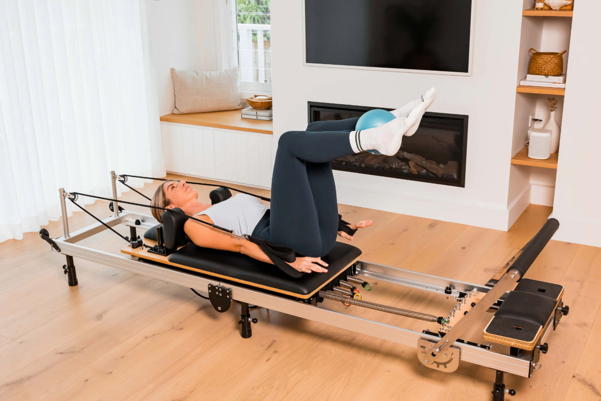 Woman using a metal and black Pilates reformer machine and pilates ball in a home setting with a fireplace and bookshelf.