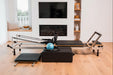 Foldable metal pilates reformer machine in a coastal style living room setting with a television and fireplace in the background. There is a black sitting box, pilates ring and blue pilates ball.