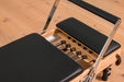 Close up of a wooden Pilates reformer machine with black cushions on a wooden floor. There are 5 colour coded springs.