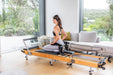 Woman using a foldable wooden pilates reformer machine in a home setting with large windows and greenery outside.
