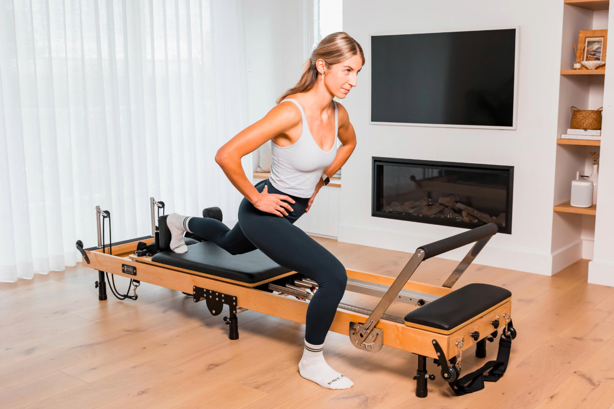 Woman exercising on a foldable wooden Pilates reformer in a home setting near the TV.