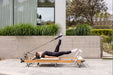 Woman using a wooden Pilates reformer machine outdoors with a modern building and greenery in the background. The reformer upholstery is off white.