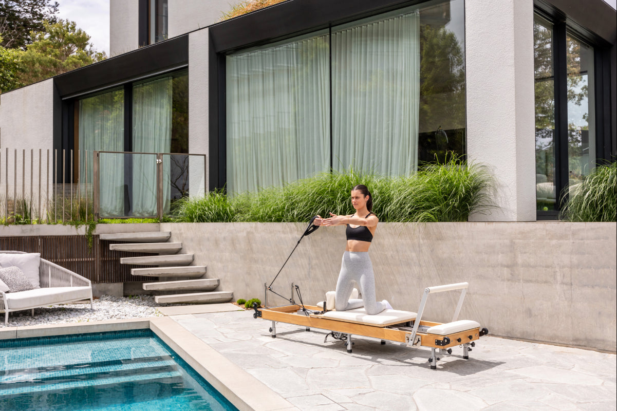 Woman exercising on a foldable wooden Pilates reformer by a poolside. The upholstery is off white.