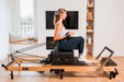 Woman using a foldable wooden Pilates reformer machine in a home setting. She is on a sitting box and pulling the leads using her arms.