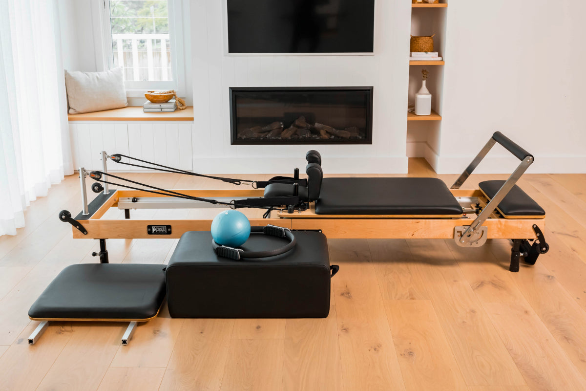 Foldable wooden reformer pilates machine in home setting near fireplace. The wood is a light colour and the upholstery is black. There is a black sitting box, pilates ring and ball as well as springboard.