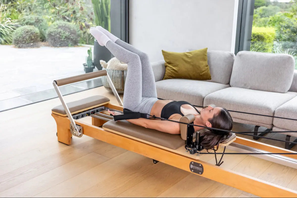 Woman using a Pioneer Pilates PP-09 Pilates reformer machine in a home setting with a couch and windows in the background.