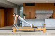 Woman exercising on a Pioneer Pilates PP-09 Pilates reformer in a modern kitchen.