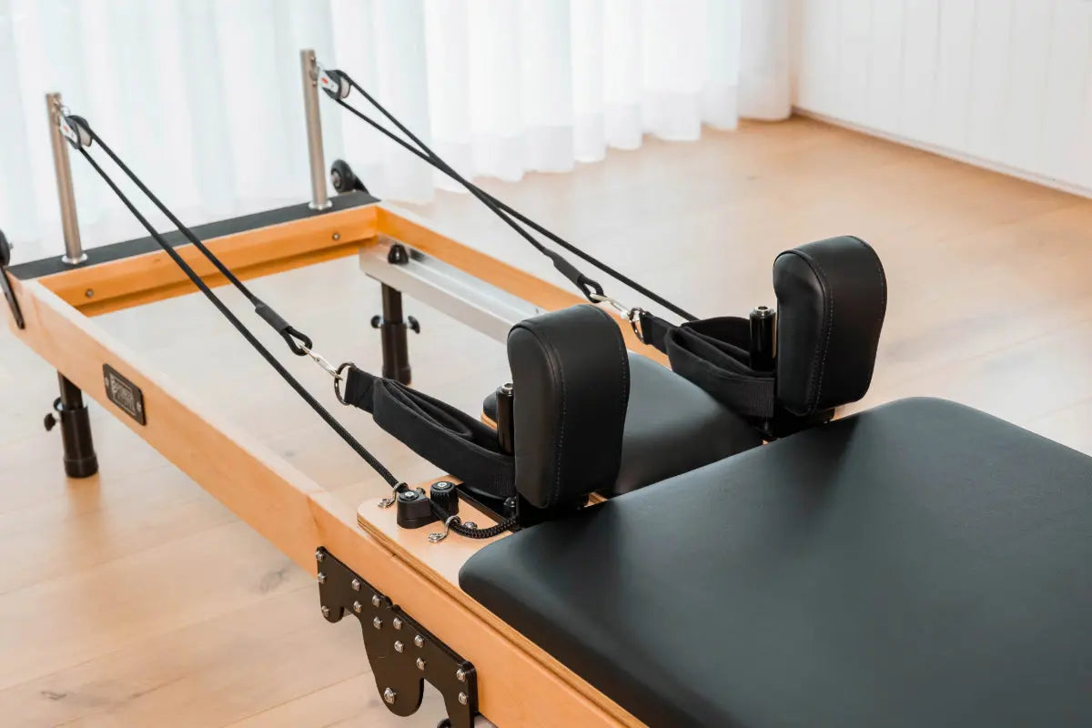 Close up of a foldable wooden Pilates reformer machine in a room with light-colored floor and white curtains. The upholstery on the reformer is black, and pulleys are black.