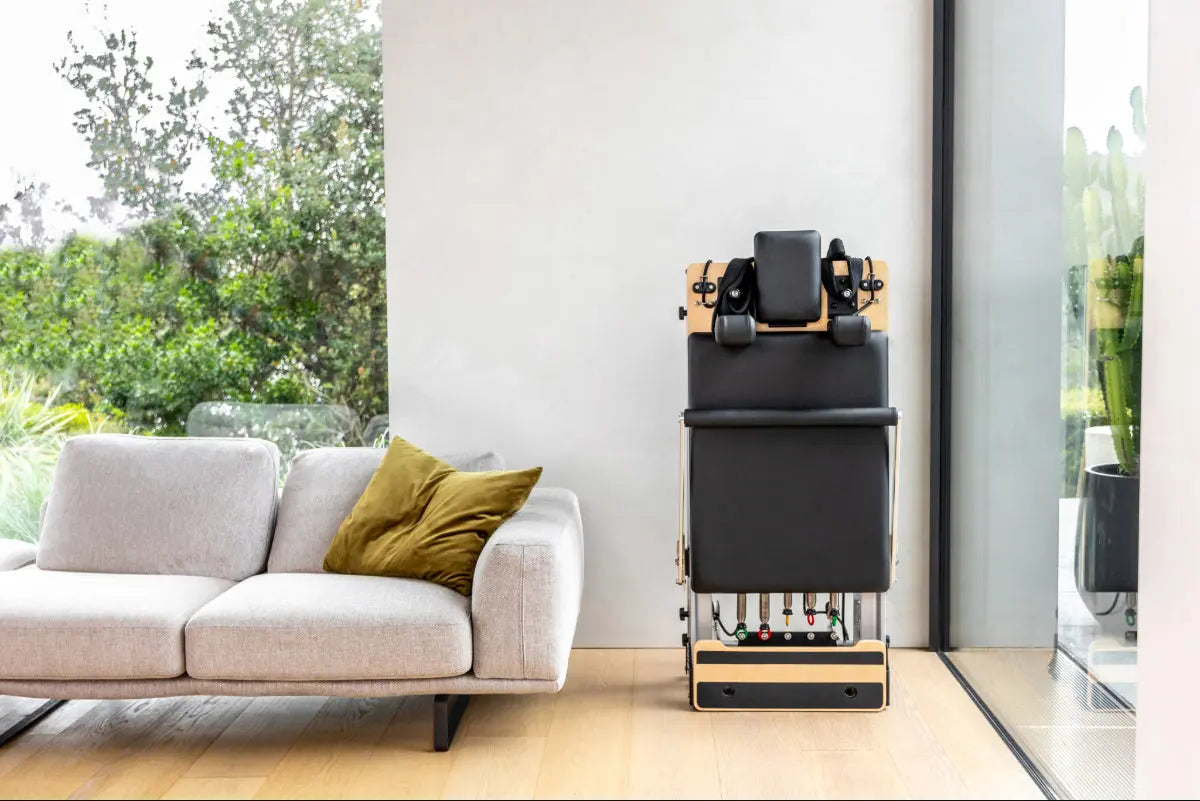 Folded reformer pilates machine in corner of modern living room. The reformer is made of metal, light coloured wood and has black upholstery.