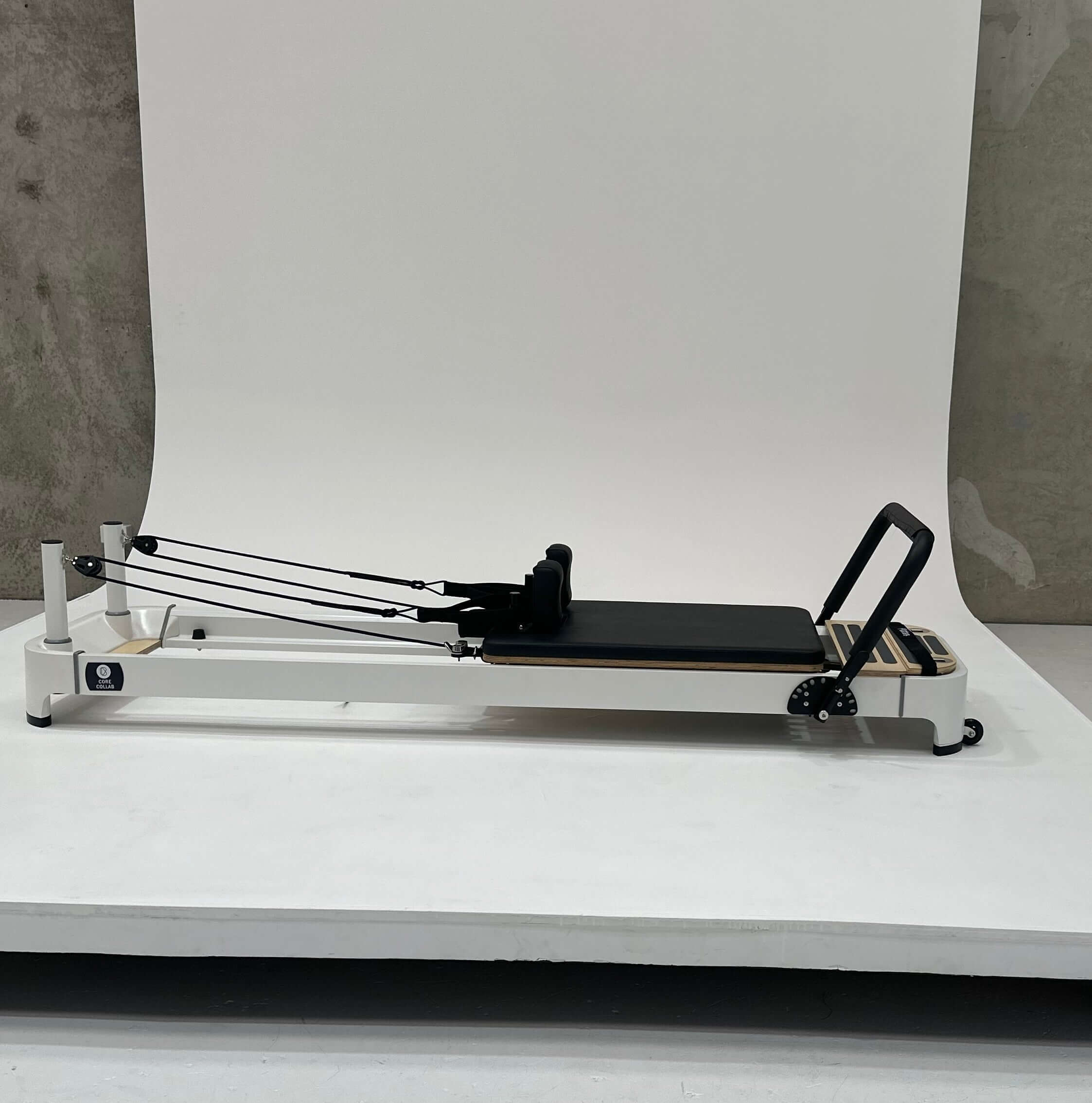 Side view of the Core Collab Queen 2.0 Pilates reformer with white frame and black upholstery in white background.