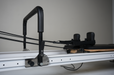 close of up the core collab queen reformer frame showing the foot bar in the middle of the white frame and the black shoulder pads and straps.