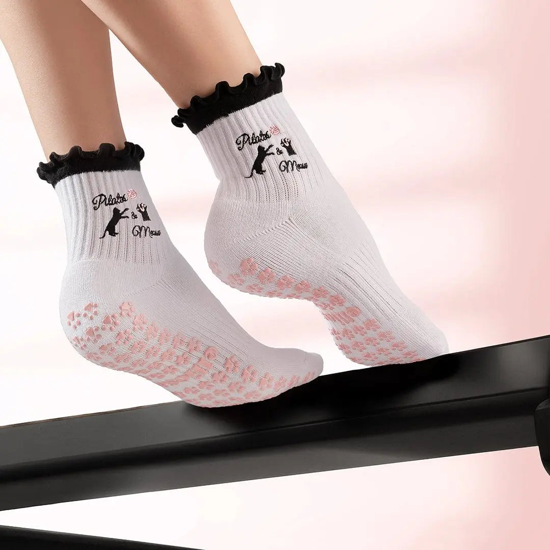 White socks with black ruffles and pink paw prints on the sole, with 'pilates & meow' text and images of cats embroidered on the ankle.