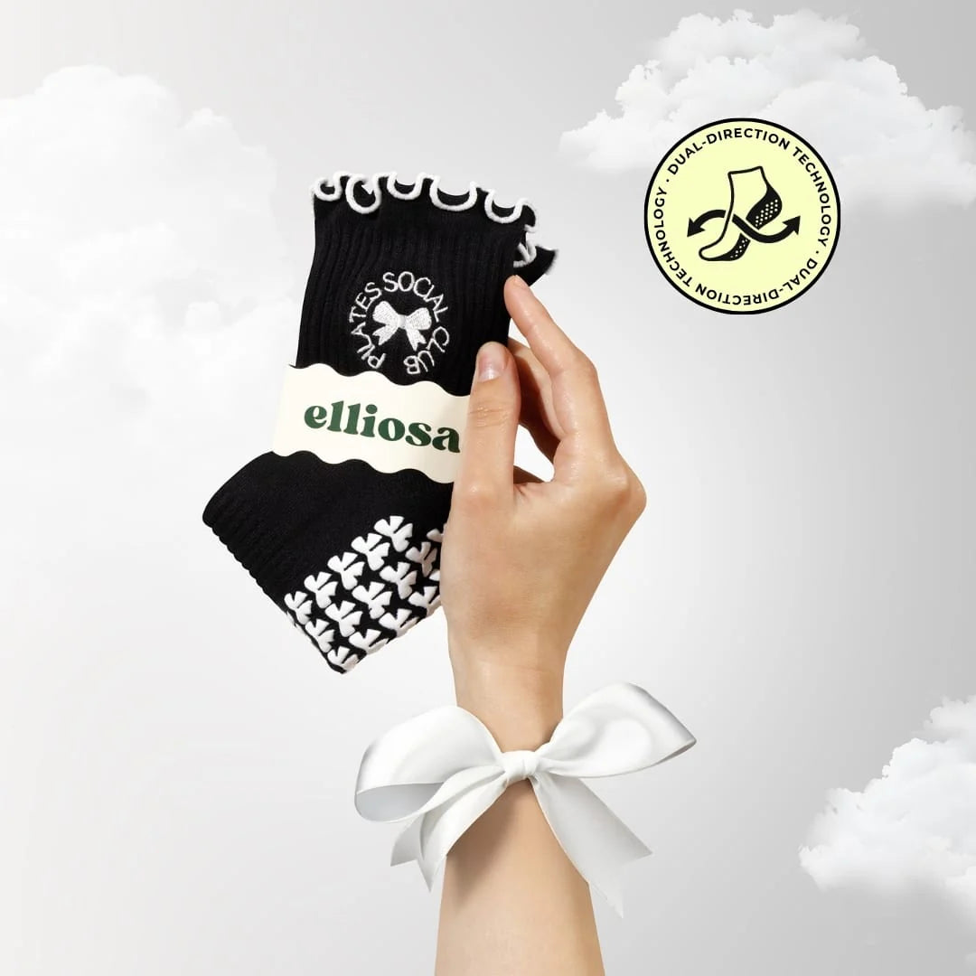 hand holding black socks with "Pilates Social Club" embroidered around a bow, with white ruffles at the ankle and white bow shaped grips on the soles.