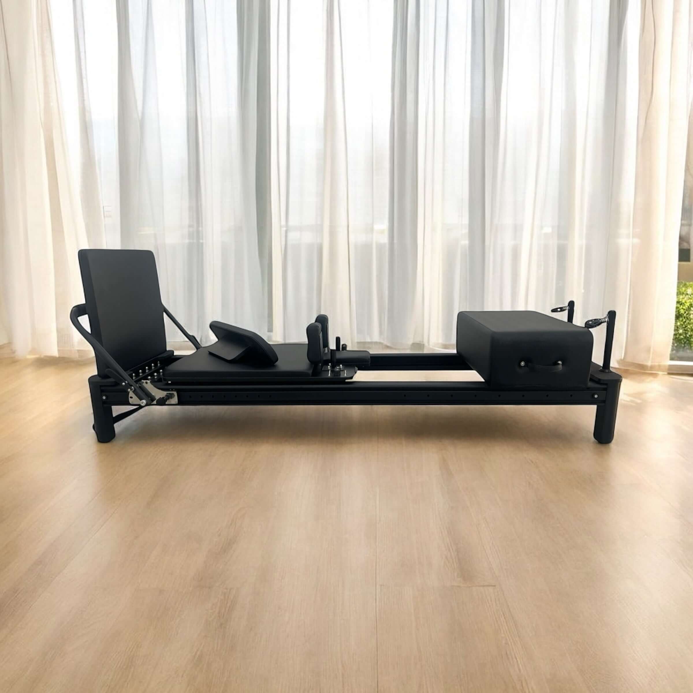 Core collab queen reformer in black frame and upholstery in lounge room with light wooden floors and sheer curtains. 